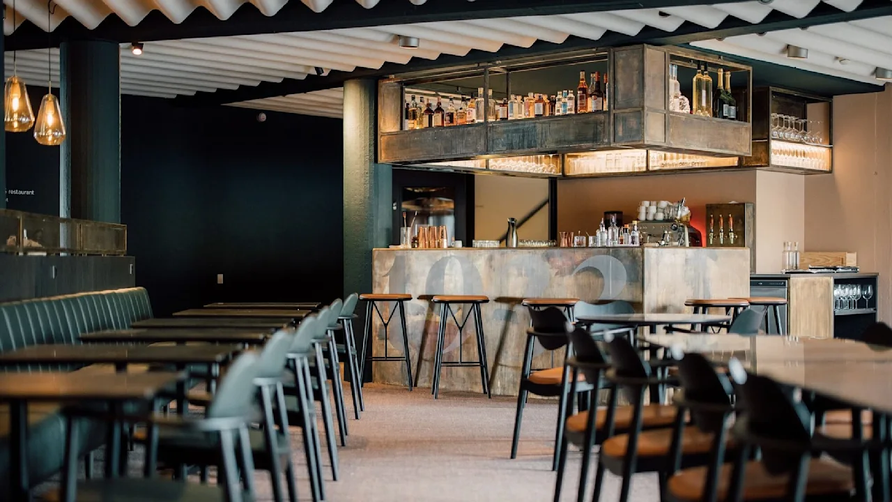 A modern bar features a sleek counter displaying various bottles. Wooden stools are arranged in front. Chairs and tables line the quiet, dimly-lit space. Visible text: "restaurant 1988".