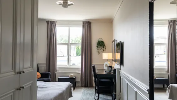 Et indbydende hotelværelse med en seng, et skrivebord og et stort vindue, der skaber en lys og behagelig atmosfære.