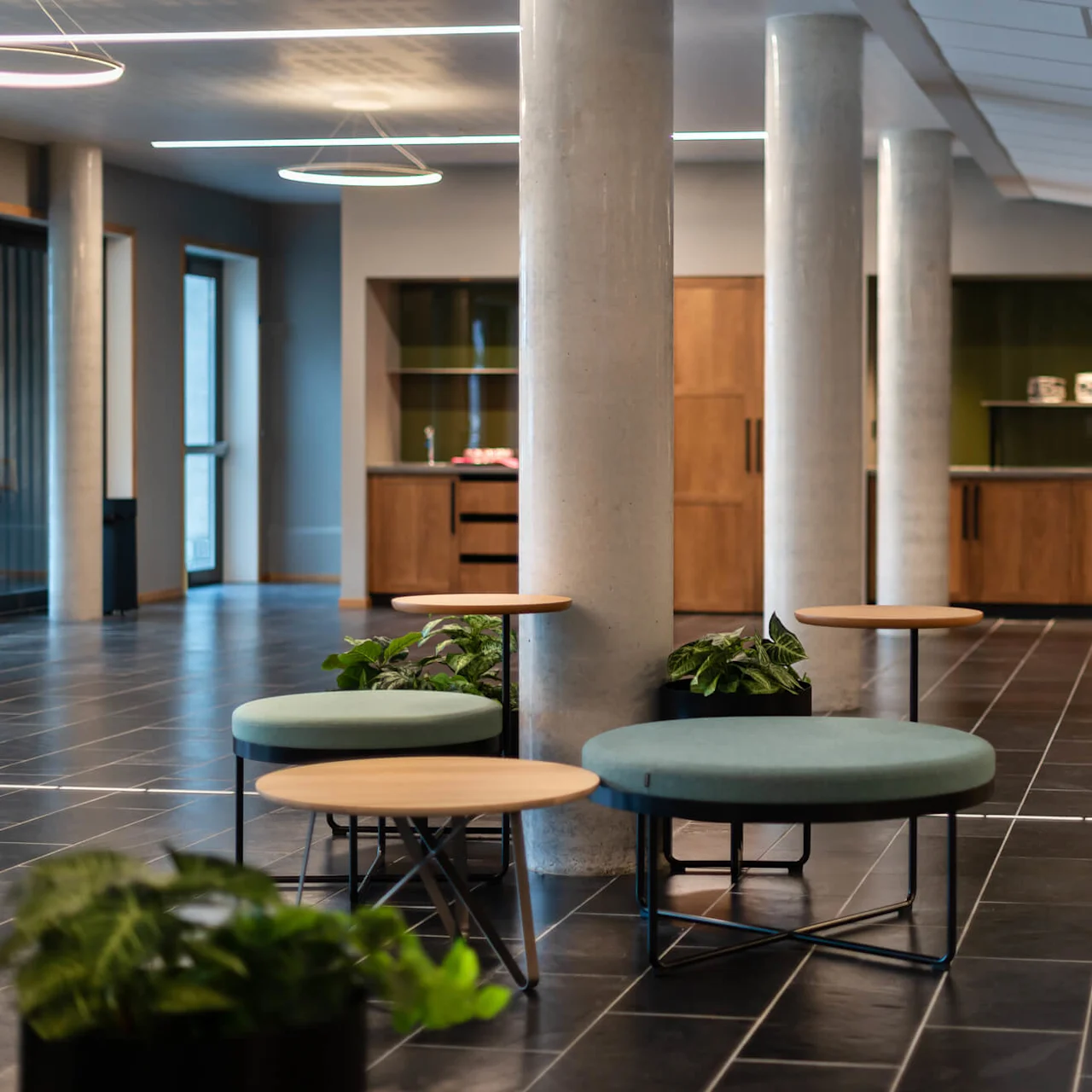 Cirkelformade sittplatser med små träbord omgivna av gröna växter i en modern, öppen yta med gråa kolonner, klinkergolv och trädskåp i bakgrunden.