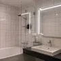 A bathroom features a bathtub with a shower, a large mirror, and a white sink on a black countertop. The walls are tiled, and there's a potted plant for decoration.