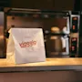 En hvit papirpose, merket "ITALIAN ON THE ROAD EST. 2025", hviler på en disk på et hotellkjøkken med en ovn.