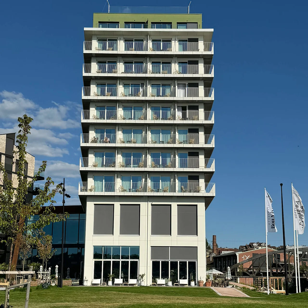 A modern, multi-story building stands tall with balconies and large windows, surrounded by a green lawn and nearby trees. Two flags read "Hotel Riviera" and "Briggen".