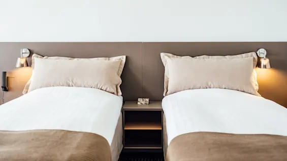 To pænt redte enkeltsenge med beige puder og sengetæpper, flankeret af vægmonterede lamper, placeret side om side i et minimalistisk rum med en lysfarvet væg.