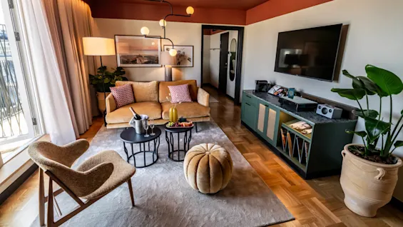 A cozy living room with a mustard sofa and decorative pillows, features a central coffee table with fruit, surrounded by a chair, pouf, and TV on the wall.