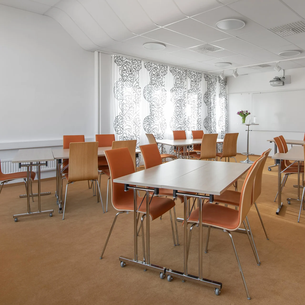 Klassrum med träbord och orange stolar arrangerade i små grupper; solljus filtrerar genom mönstrade gardiner, rummet är upplyst med beige matta. En whiteboard och projektor är synliga.