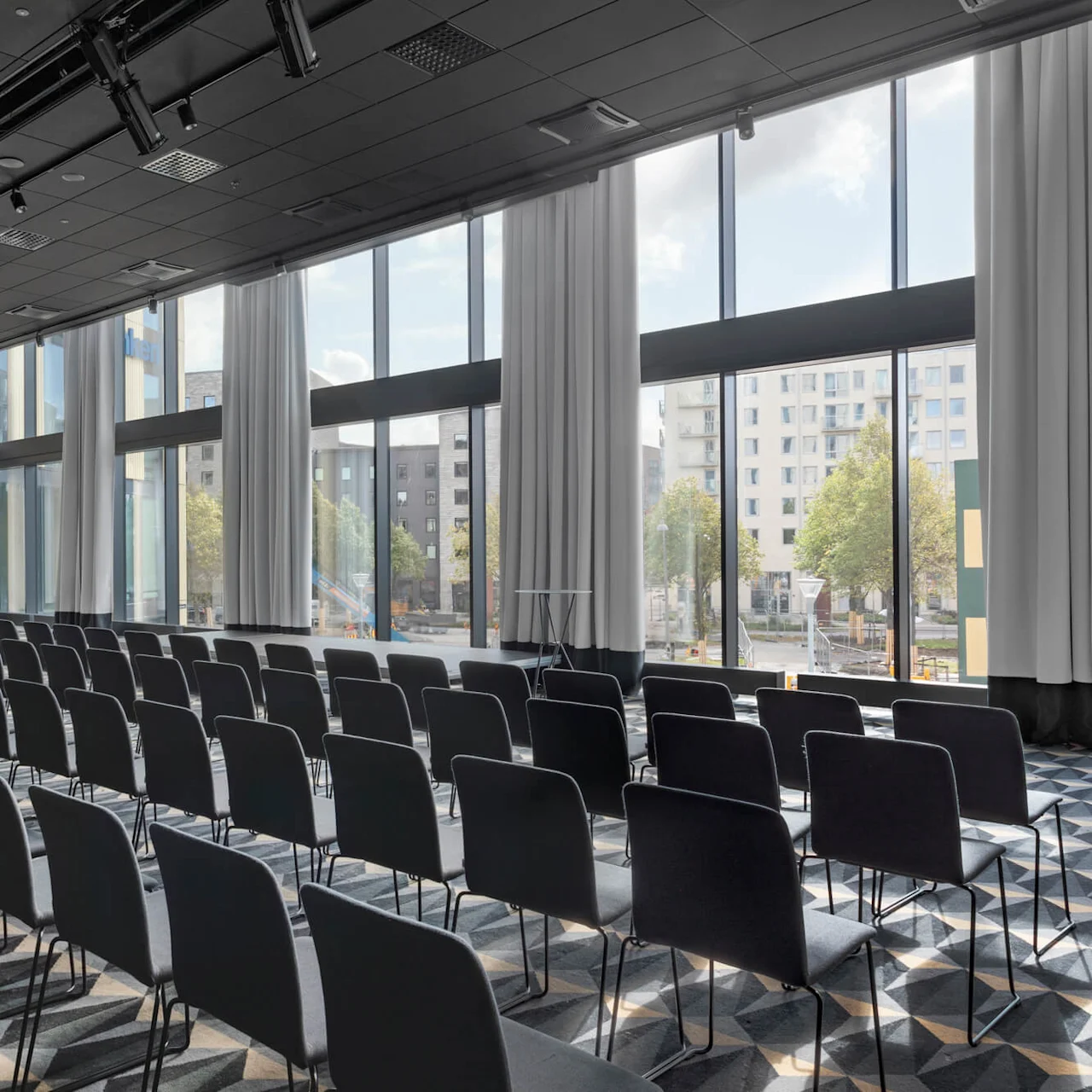 Rader av tomma svarta stolar är prydligt arrangerade i ett konferensrum med stora fönster som släpper in naturligt ljus på den geometriskt mönstrade mattgången.