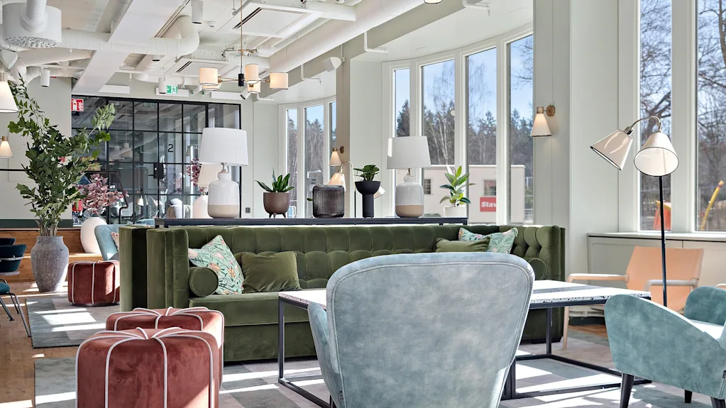 Lyst og moderne hotellobbyområde med en grønn fløyelsofa, lenestoler og store vinduer som slipper inn sollys.
