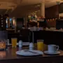 Et bord dekket med bestikk, juice og kaffe med utsikt over en svakt opplyst restaurant. I nærheten har en buffet forskjellige retter. En dekorativ lysestake gir omgivende belysning.