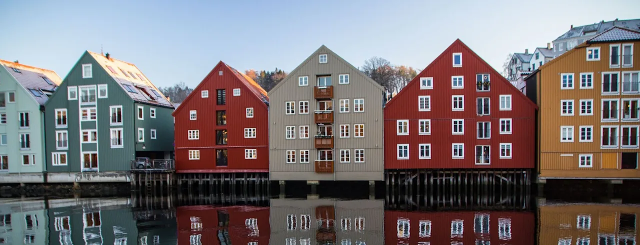 Farverige bygninger ved vandet med gavlhuse reflekteret i roligt vand under en klar himmel.