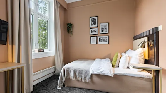 Et koselig hotellrom med en komfortabel seng, et stort vindu og innrammet kunst på veggen, som skaper en innbydende atmosfære.