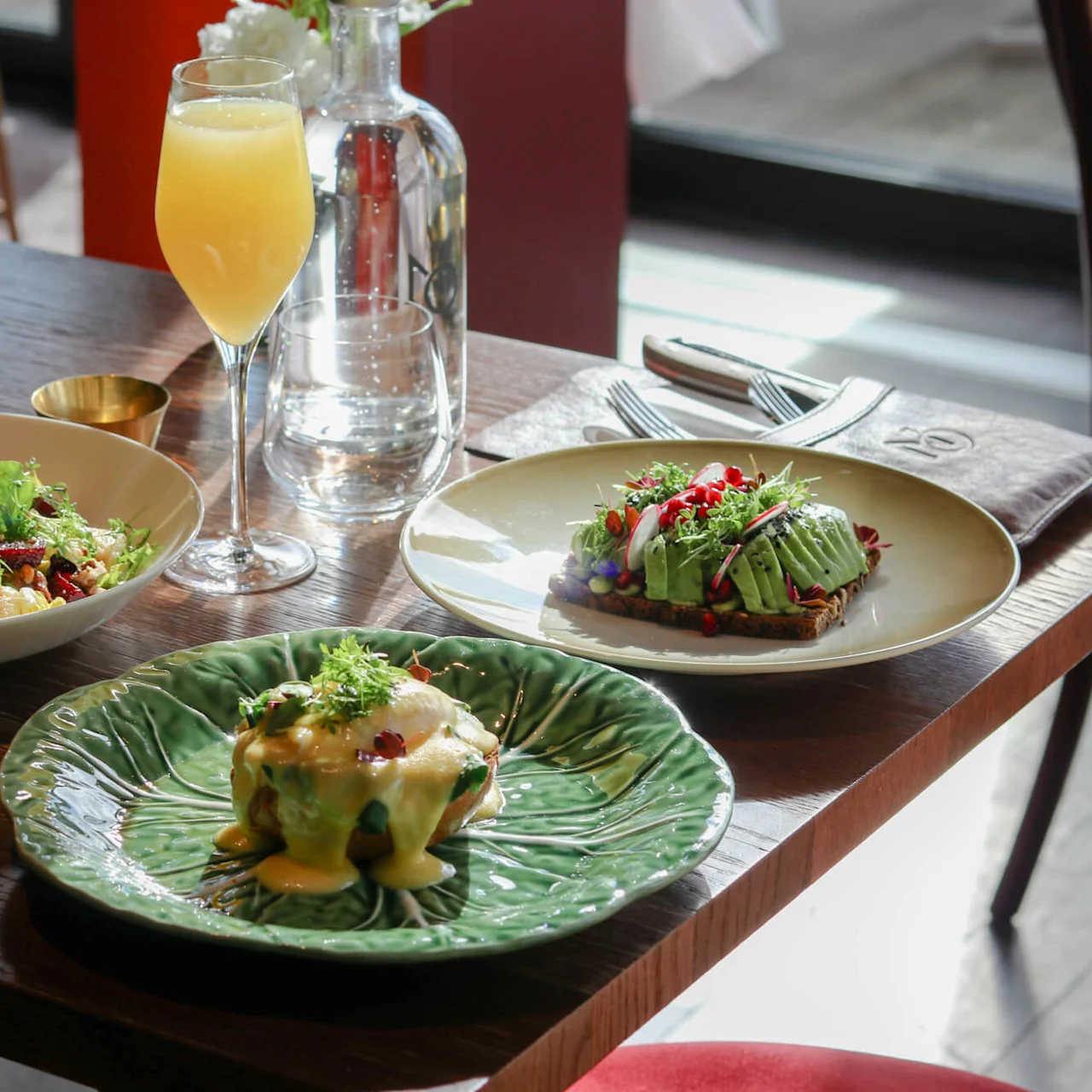 Three plates of gourmet food and a glass of orange beverage sit on a wooden table near a window. Utensils are neatly placed on a folded napkin.