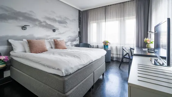 A spacious bedroom features a double bed with white bedding and brown pillows, flanked by nightstands with flowers; natural light filters through sheer curtains, illuminating dark floors.