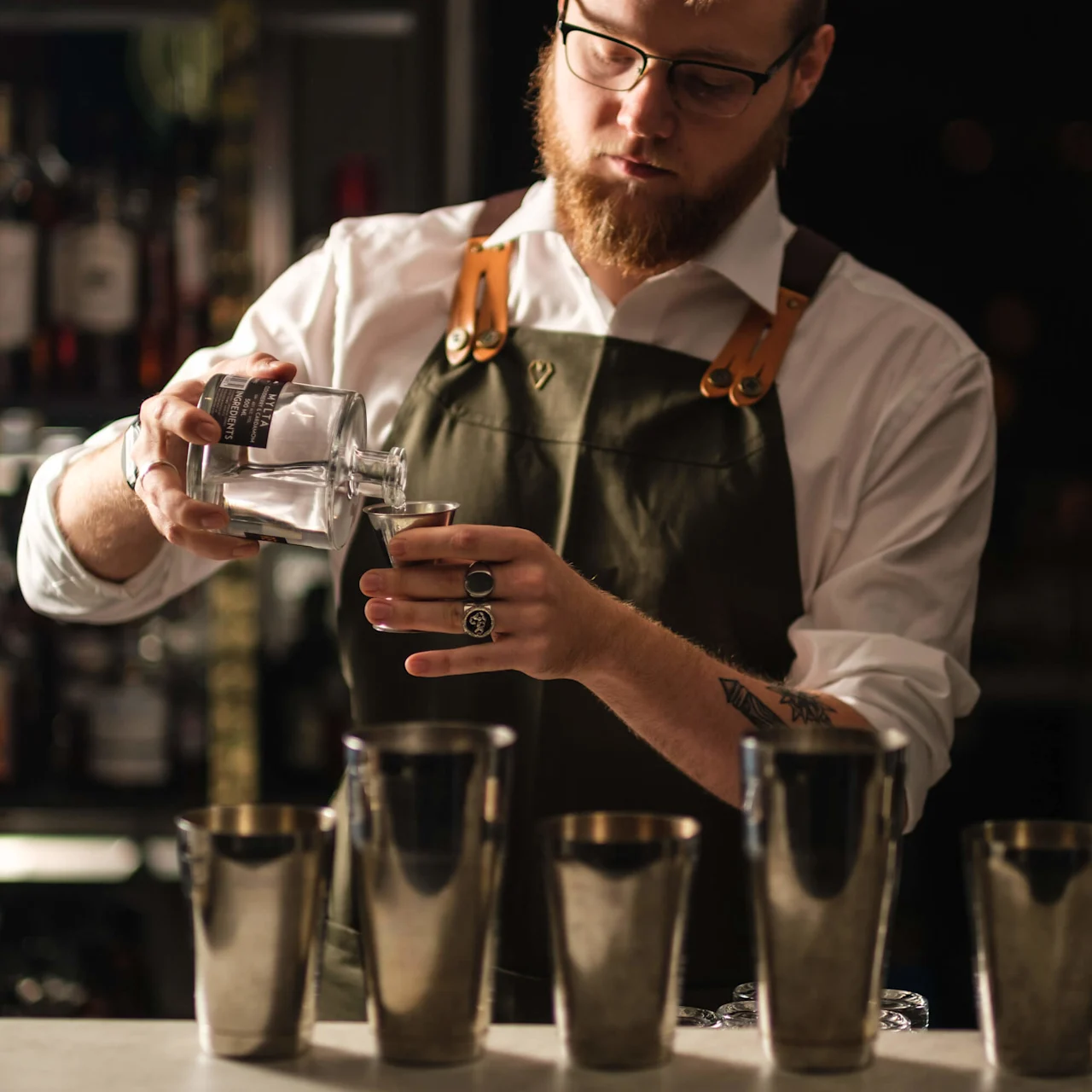 Bartender häller vätska från en flaska i mätglas, bär en vit skjorta och grön förkläde i en bar med flaskor på hyllor bakom.