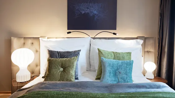 Elegant bed with plush green and blue cushions, set in a cozy bedroom. Ambient lighting from two white lamps on bedside tables, with a dark abstract painting hanging above.