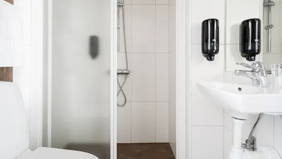 Dusch med frostat glasdörr, intill en toalett och handfat. Svarta dispensrar är monterade på en vitkaklad vägg ovanför en modern kran. Vitt handduk hänger på sidan.
