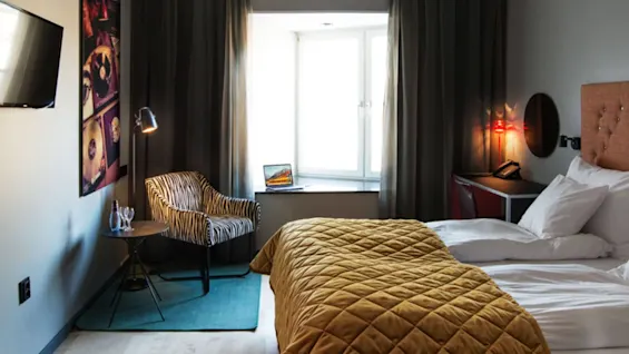 Ett mysigt hotellrum med en säng med senapsgult täcke, zebrarandig stol med bord och en laptop på fönsterbrädan. Inramade skivor och en väggmonterad TV pryder väggarna.