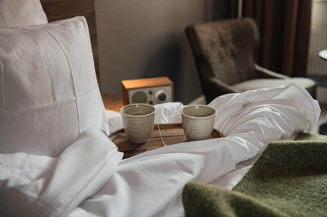 Ceramic cups sit on a wooden tray atop a rumpled, white bed. Nearby, a brown chair and a small wooden speaker add to the cozy atmosphere.