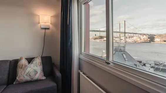 A lit wall lamp casts a warm glow above a patterned pillow on a gray sofa, beside a window overlooking a bridge and waterfront with boats.