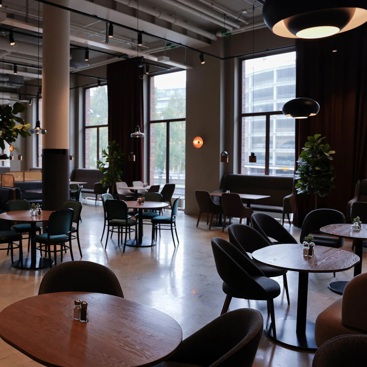 Modern café interior with empty chairs and tables, large windows, industrial ceiling, and plants, suggesting a comfortable urban dining space.