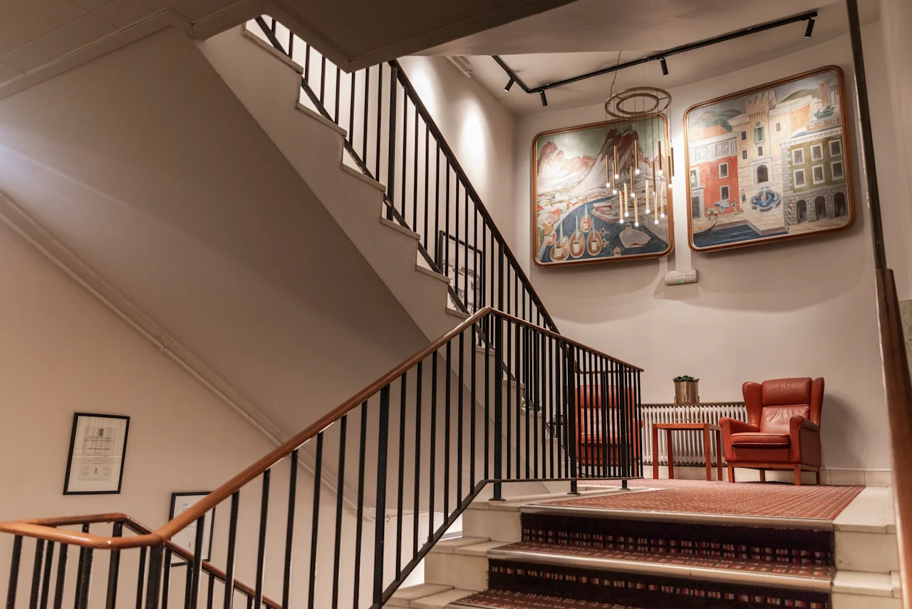 Elegant staircase with red carpet, leading to a cozy seating area with red armchairs and artwork at Home Hotel Grand Bodø.