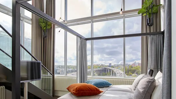 En moderne seng med fargerike puter står under et stort vindu, med utsikt over byen. En TV er montert i nærheten. Høye gardiner rammer inn loftstilen.