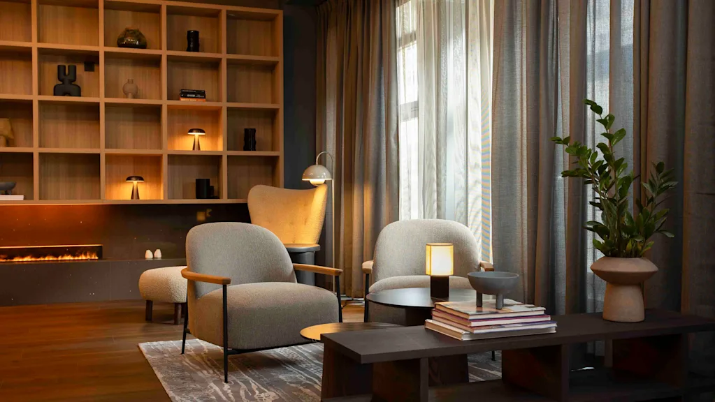 Two armchairs sit beside a wooden coffee table with books and a lamp, in a cozy living room featuring a lit fireplace, shelves with decor, and large, curtained windows.