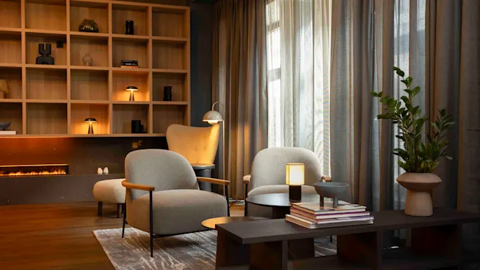 Kaksi nojatuolia on puisen sohvapöydän ääressä, pöydällä kirjoja ja lamppu, viihtyisässä olohuoneessa, jossa on takka, hyllyillä koriste-esineitä ja suuret verholliset ikkunat.