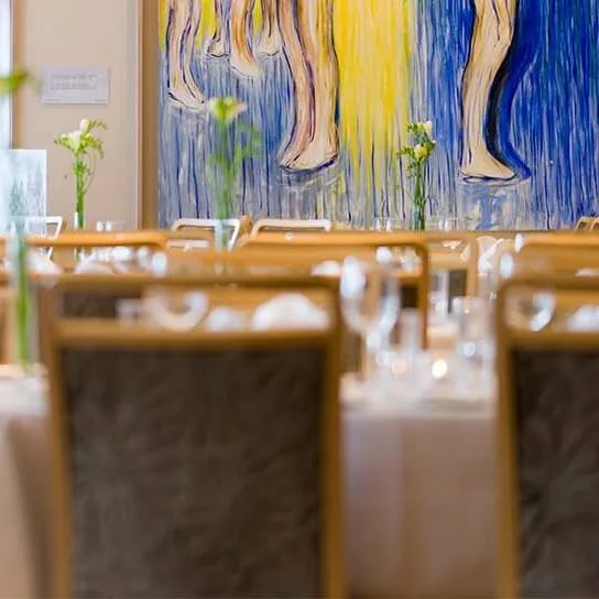 Elegante spiseborde dækket med glas og blomster, placeret i et rum med et stort abstrakt maleri, der viser ben i blå og gule nuancer på væggen.