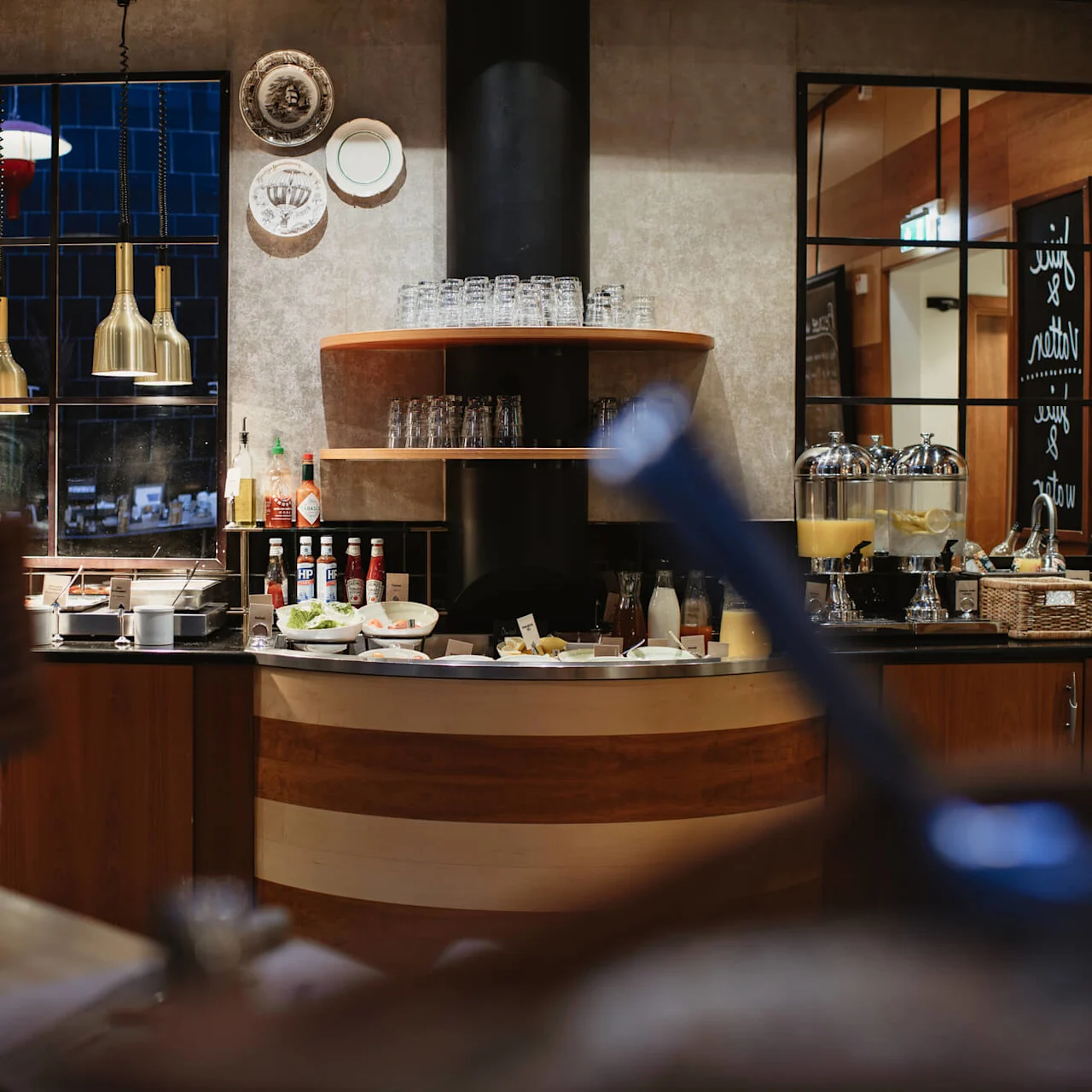 A kitchen counter adorned with condiments and food items. Above, golden pendant lights hang. In the background, glassware and decorative plates are displayed. Text: "Juice & water".