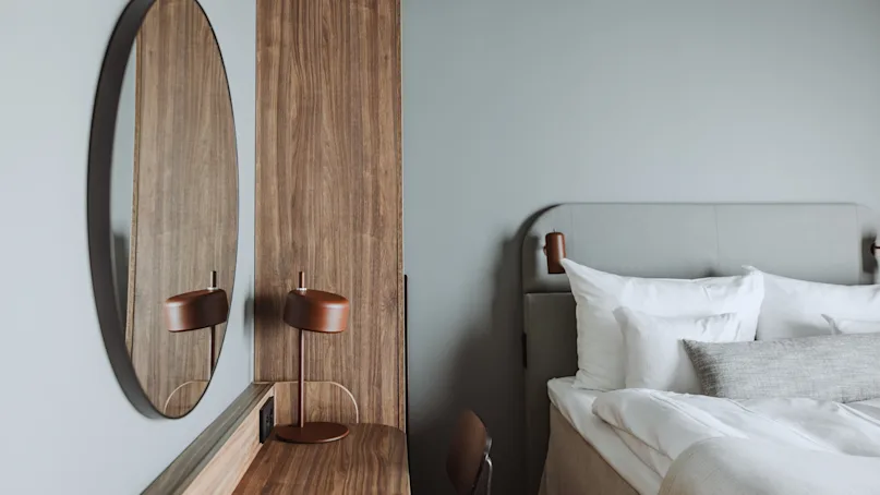 Round mirror reflecting a minimalist lamp on a wooden desk. Modern bedroom with a soft gray bed and white bedding nearby.