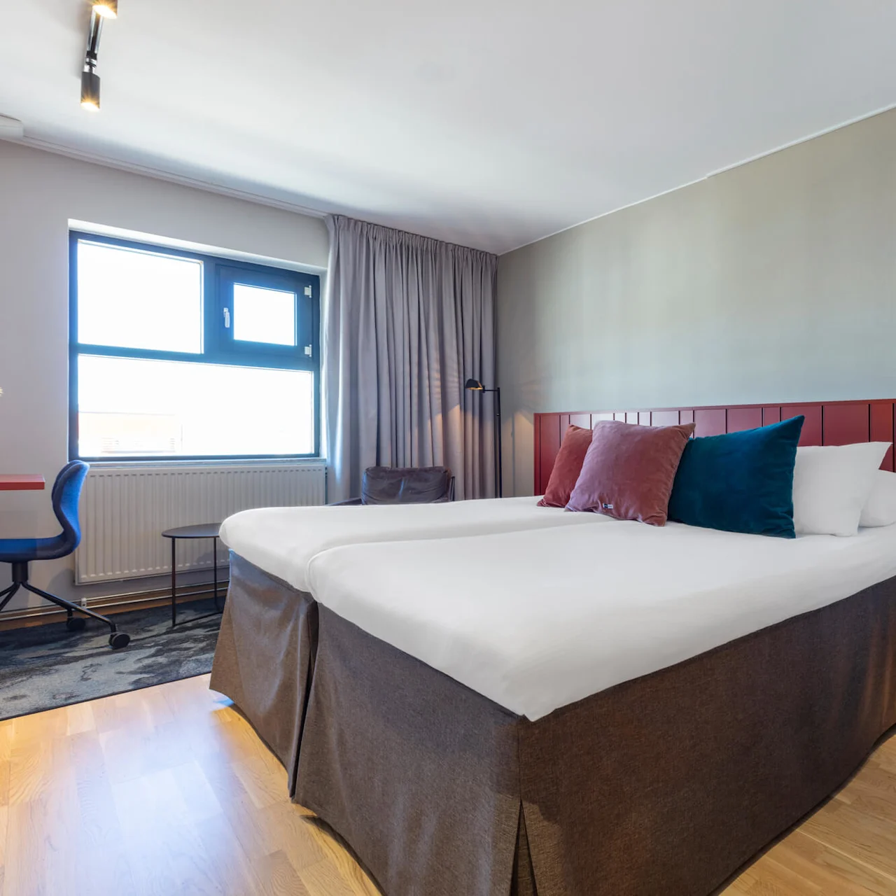 A neatly made bed dominates a modern hotel room with simple furniture, artwork on the wall, and daylight streaming through a window.