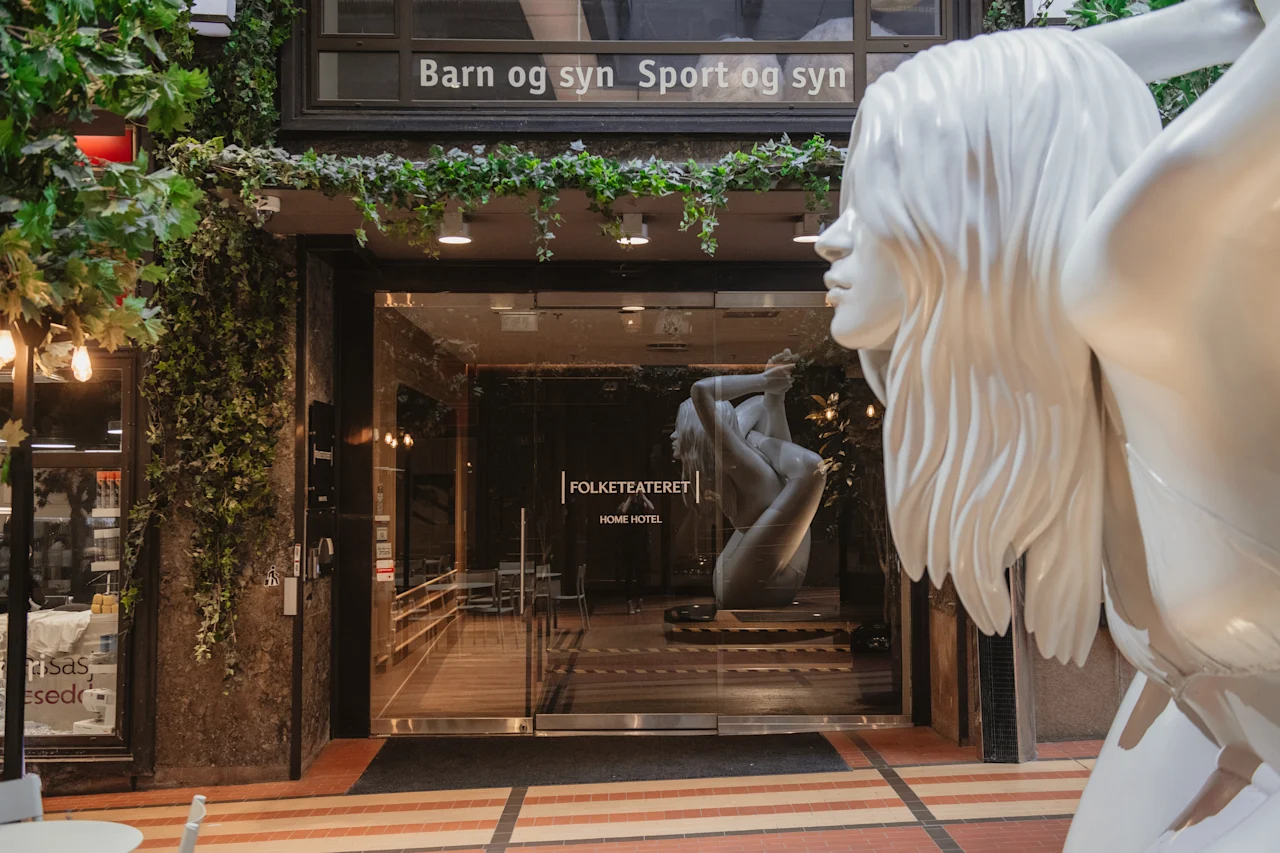 Building entrance with green plants. Inside, a statue in a yoga pose. Text above door: Barn og syn Sport og syn.