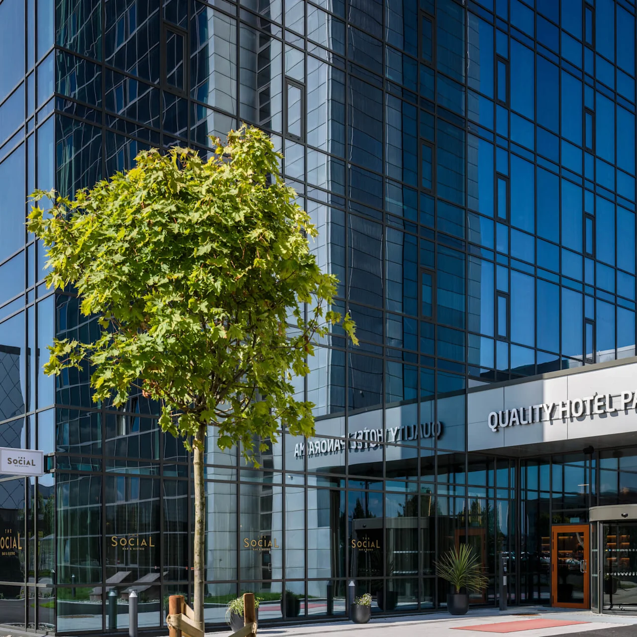 En moderne bygning i glass har et tre i forgrunnen. Inngangen viser teksten "QUALITY HOTEL PANORAMA" og "The Social Bar & Bistro" under en klar himmel.