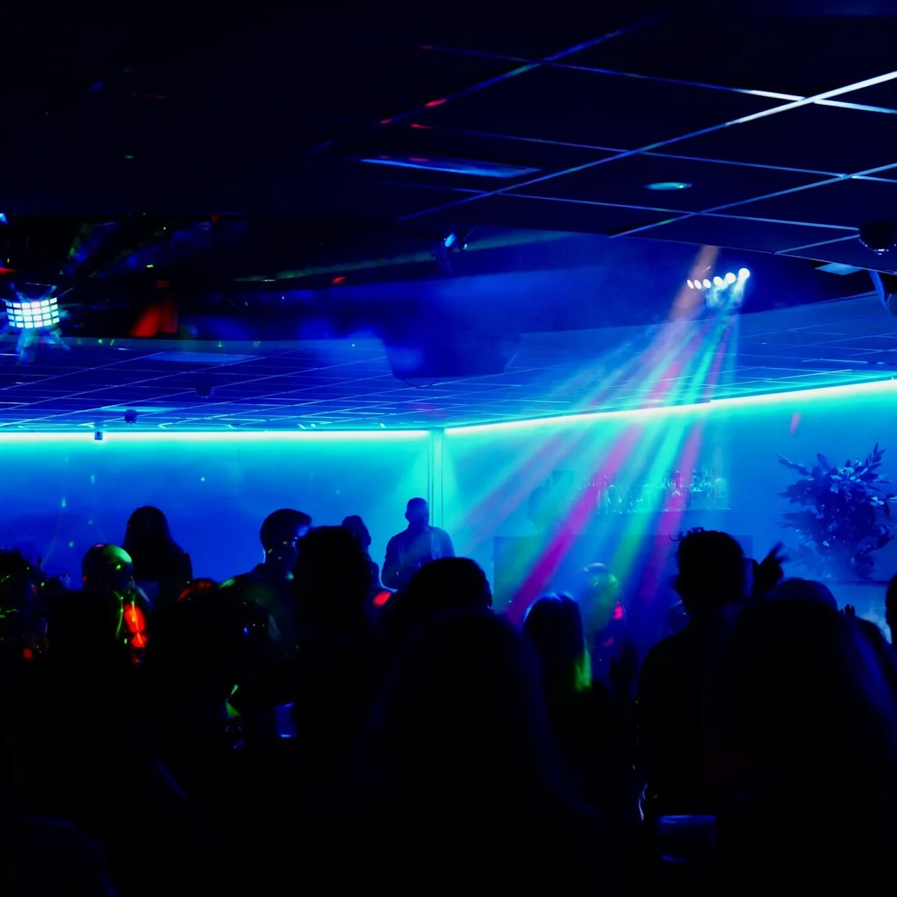Silhuetpersoner danser under farverige lys i en klub. En DJ står i baggrunden, omgivet af livlig blå og pink belysning, der skaber en livlig atmosfære.