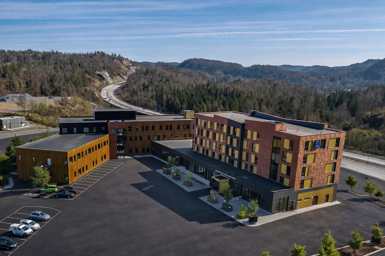 Ett modernt hotellkomplex med parkering, omgivet av skogsklädda kullar och en motorväg i bakgrunden. Synlig text: Hotell.