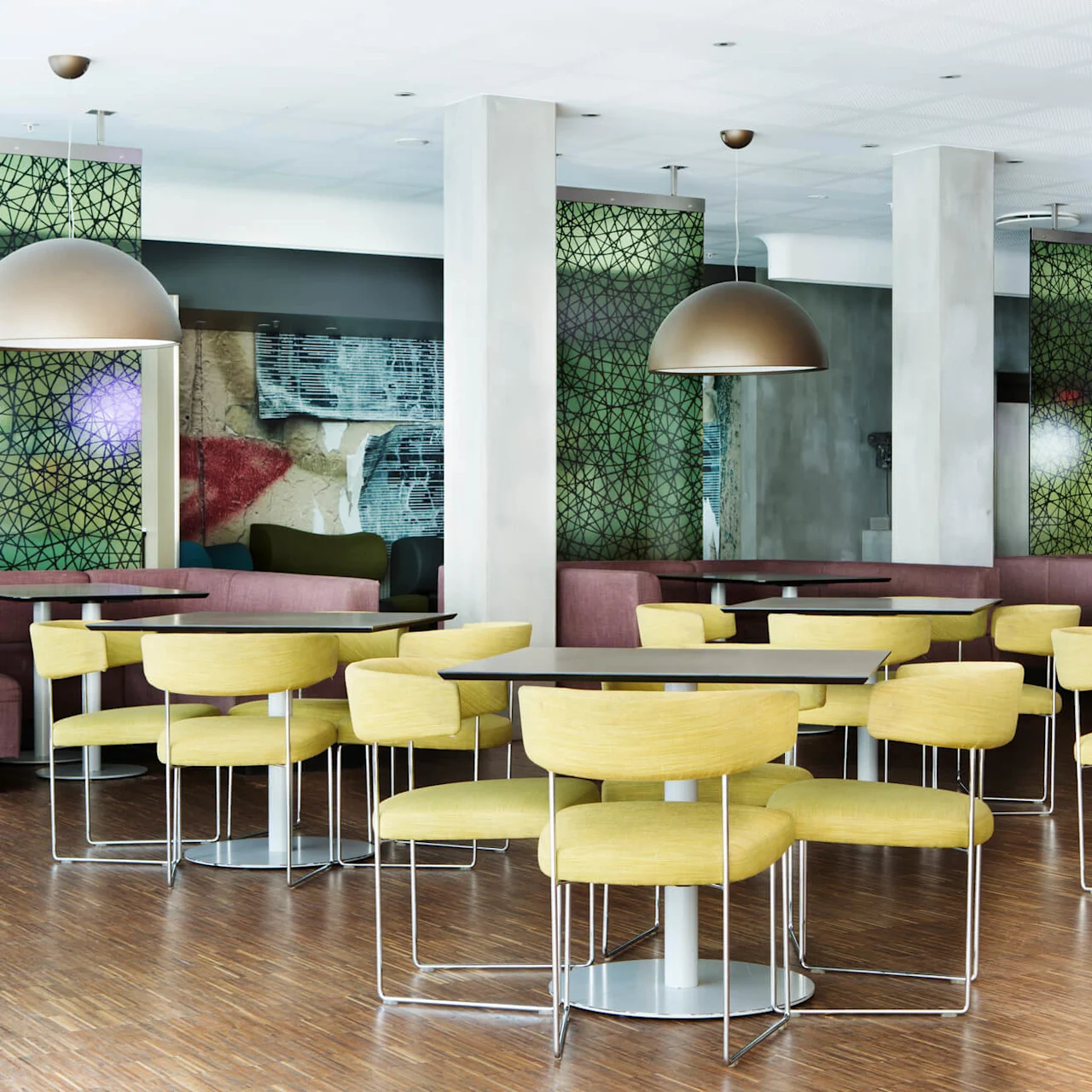 Yellow chairs are arranged around tables in a brightly lit, modern dining area with decorative green panels and large hanging lamps.