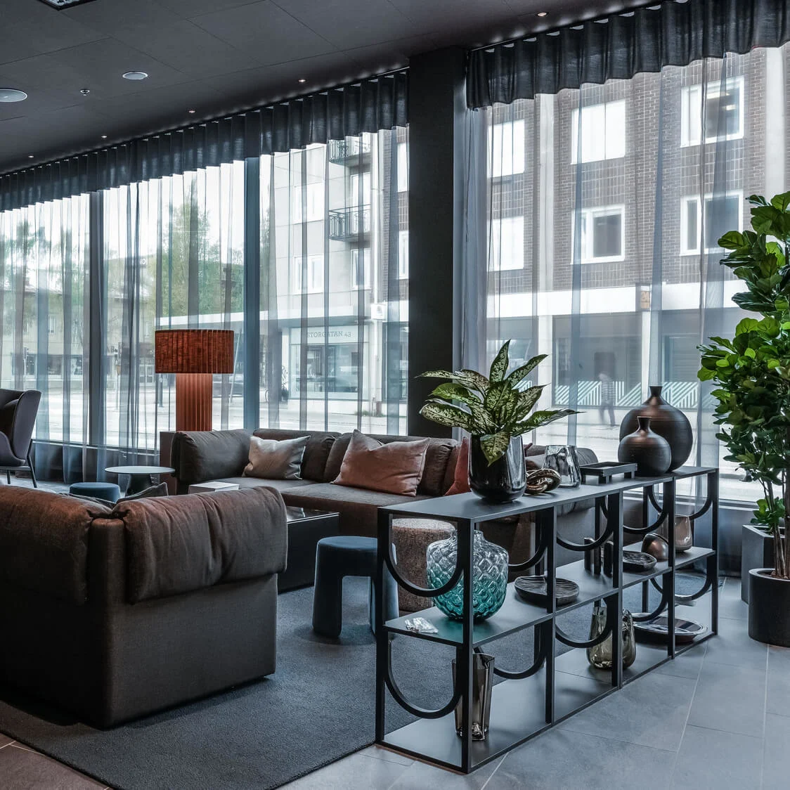 Moderne lounge med plyssofaer og stole, omgivet af planter. Store vinduer filtrerer naturligt lys, hvilket forbedrer den sofistikerede atmosfære. Hylder viser dekorative genstande, der skaber et indbydende miljø.