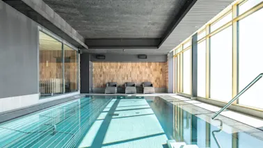 En inomhuspool med klart vatten ligger bredvid tre gråa solstolar. Solljus filtreras genom stora fönster till höger och ger rummet ett avslappnande ljus.