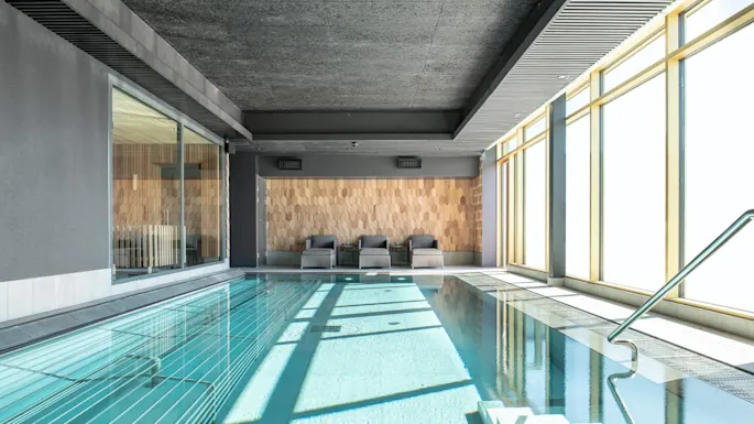 Et innendørs basseng med klart vann ligger ved siden av tre grå liggestoler. Sollys filtrerer gjennom store vinduer til høyre, som lyser opp rommet med en avslappende atmosfære.