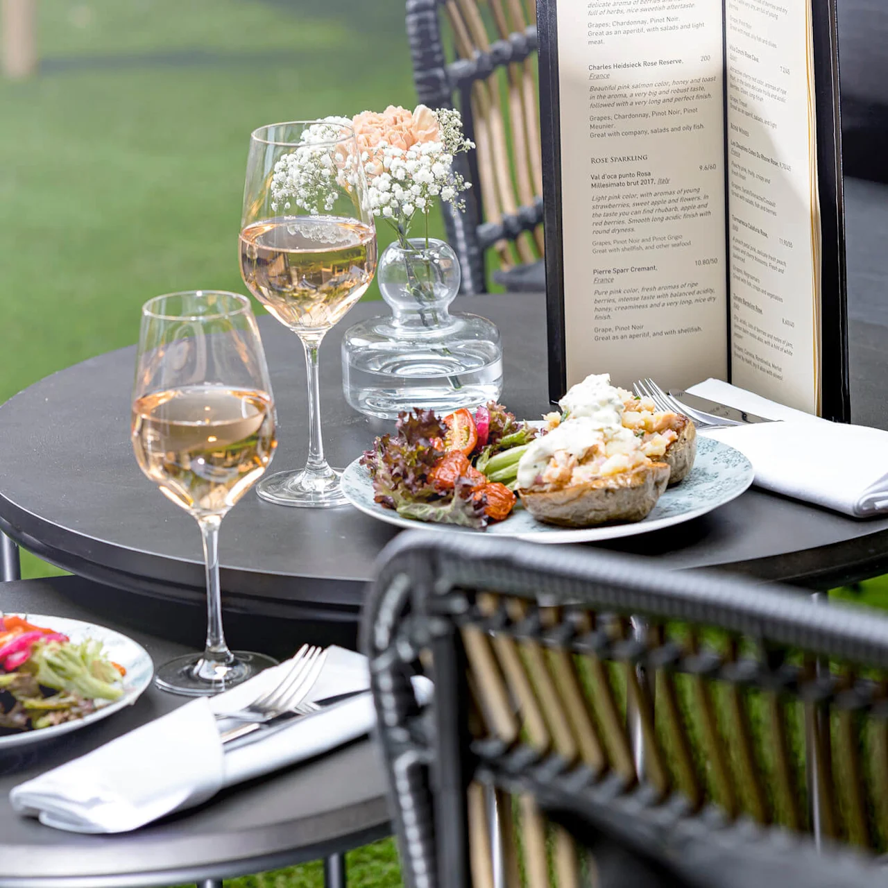 Two elegant dishes with salads and two glasses of rosé wine sit on a garden table. A menu and small flower vase add to the outdoor dining experience.