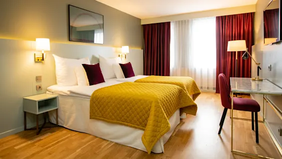 Ett mysigt hotellrum med dubbelsängar täckta av gula täcken, kompletterade med vinröda gardiner. Ett skrivbord med stol och lampa står vid fönstret, under mjuk belysning.