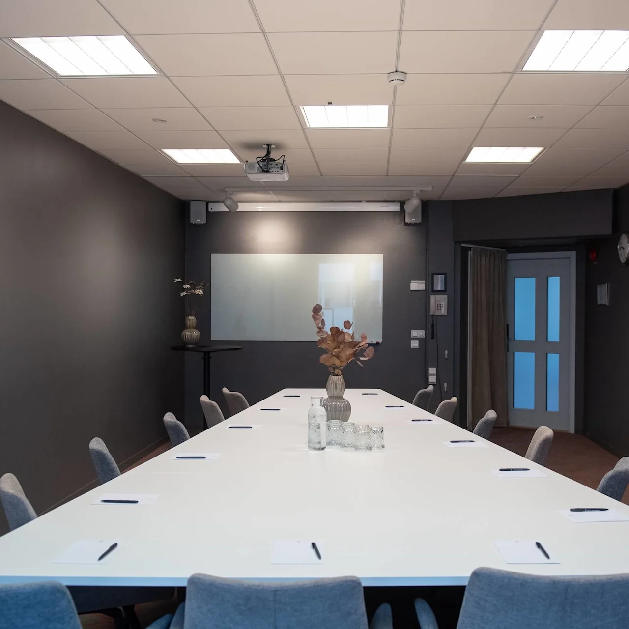 Long white conference table with gray chairs, notepads, and pens, situated in a modern room with dark walls and ceiling lights. A decorative plant sits in the center of the table.