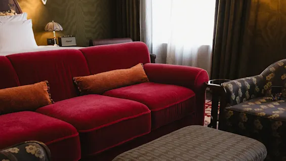 Et koselig romhjørne med en rød sofa, mønstret lenestol, fotskammel og et tent taklampe som reflekterer på et blankt bord.