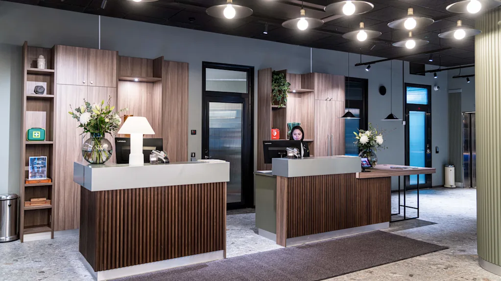 Resepsjonsdisker med trepaneler i en moderne lobby. En person ved en disk jobber på en datamaskin. Overhengende lys opplyser rommet, dekorert med blomster og en lampe.