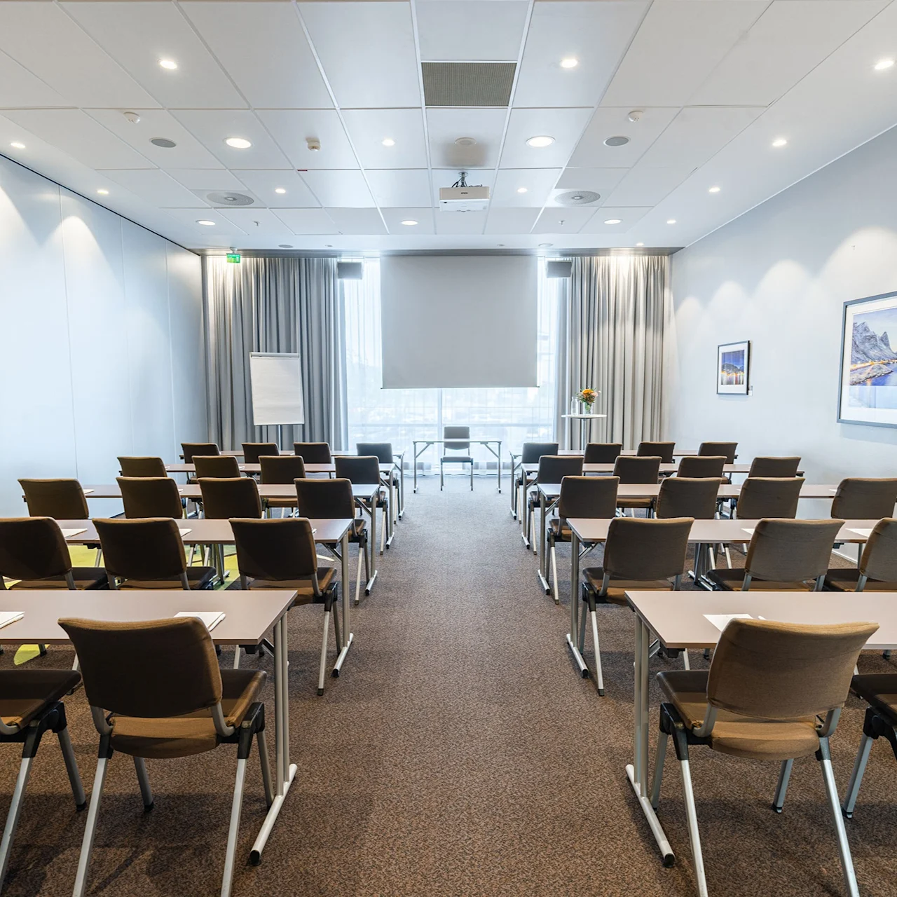Rows of empty chairs face a projector screen in a bright conference room. The setting has tables with notepads, beige walls, and framed pictures.