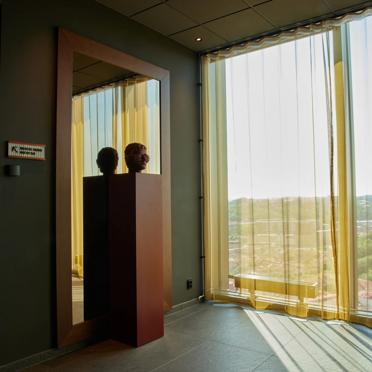 Byster är placerade på en piedestal och reflekteras i en hög spegel. Solljus strömmar genom stora fönster med skira gardiner. En skylt på väggen lyder "K konferens konferensrum."
