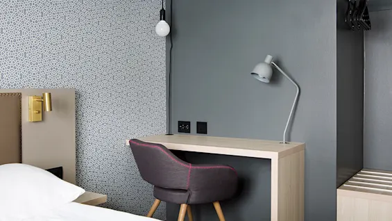 A modern desk with an adjustable lamp, positioned in a bedroom with patterned wallpaper and a bed partially visible.