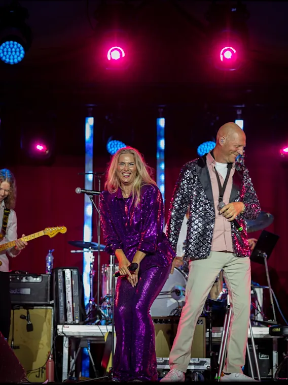 Et livlig band opptrer på scenen under sterke lys, med musikere som spiller gitar og sangere som smiler og engasjerer publikum.