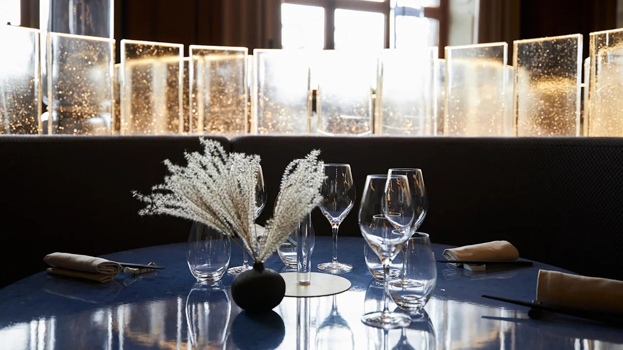 Bord dekket med vinglass og en svart vase med hvite tørkede blomster. I bakgrunnen fanger belyste glasspaneler sollyset og skaper en varm atmosfære.