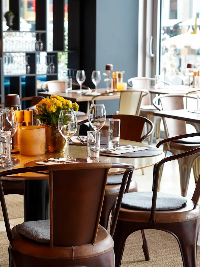 A set table with flowers and glassware, arranged on wooden chairs in a cozy, warmly lit restaurant, with large windows and decorative vases visible in the background.